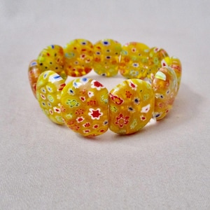 May include: A yellow bracelet with a floral design. The bracelet is made of oval-shaped beads with a yellow base and colorful floral patterns in red, white, blue, and yellow. The bracelet is elastic and stretches to fit the wrist.