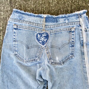 May include: A pair of light blue denim jeans with a white heart-shaped patch with a floral design on the back pocket. The jeans have a zipper on the side seam.