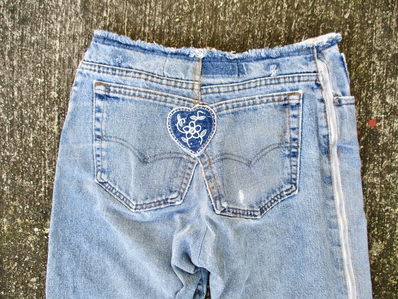 May include: A pair of light blue denim jeans with a white heart-shaped patch with a floral design on the back pocket. The jeans have a zipper on the side seam.