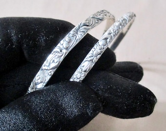 May include: Two silver bangles with intricate floral and foliate designs. The bracelets are detailed with embossed patterns of flowers, leaves, and other natural elements. The bangles are displayed against a black background.