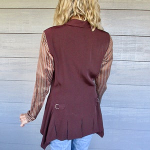 May include: A brown sleeveless vest with a long, asymmetrical hemline. The vest has a collar and a button closure. The vest is worn over a striped shirt.