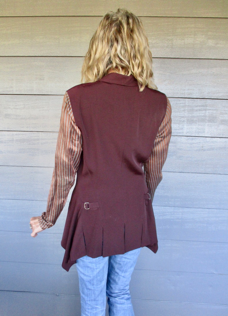 May include: A brown sleeveless vest with a long, asymmetrical hemline. The vest has a collar and a button closure. The vest is worn over a striped shirt.