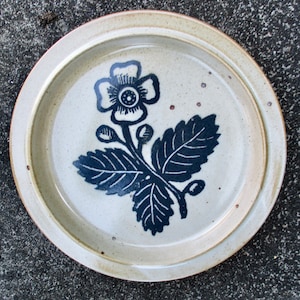 May include: A white ceramic plate with a blue and white floral design. The design features a flower with five petals and leaves.