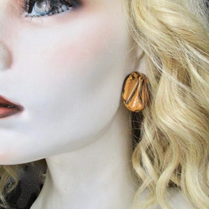 May include: A close-up of a mannequin's face and shoulder, with a focus on a single gold-colored earring. The earring is an oval shape with a textured, layered design. The mannequin has blonde hair and is wearing makeup.