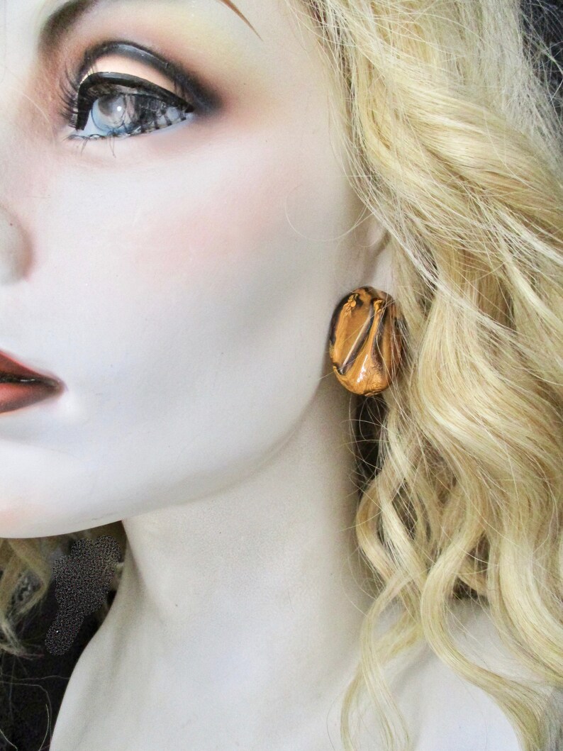 May include: A close-up of a mannequin's face and shoulder, with a focus on a single gold-colored earring. The earring is an oval shape with a textured, layered design. The mannequin has blonde hair and is wearing makeup.