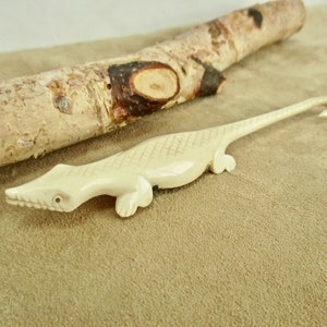 May include: A white carved bone lizard with a long tail and a hook on the end. The lizard is lying on a brown surface.
