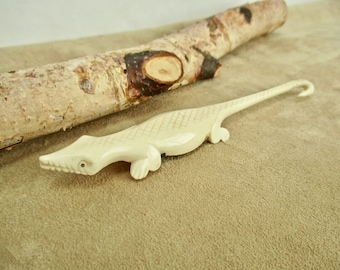 May include: A white carved bone lizard with a long tail and a hook on the end. The lizard is lying on a brown surface.