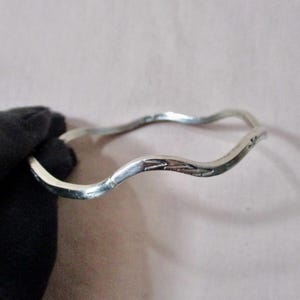 May include: A silver-colored bangle bracelet with a wavy design. The bracelet has a smooth, polished finish and a subtle, engraved pattern on one section. The bracelet is approximately 2.5 inches in diameter.