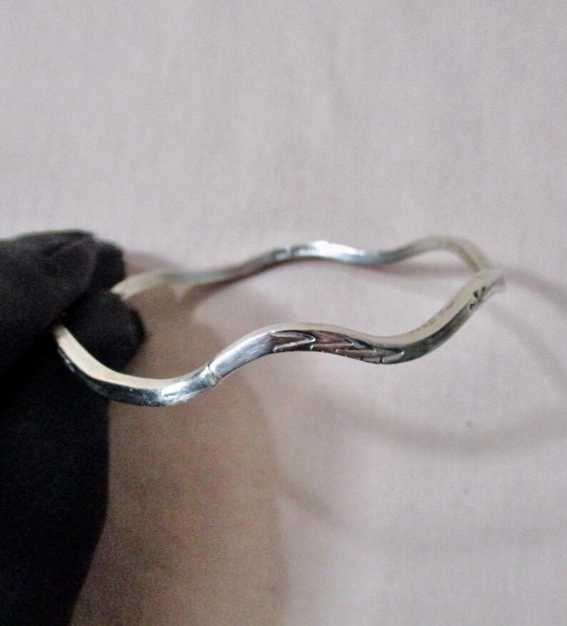 May include: A silver-colored bangle bracelet with a wavy design. The bracelet has a smooth, polished finish and a subtle, engraved pattern on one section. The bracelet is approximately 2.5 inches in diameter.