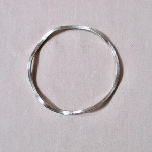 May include: A silver-colored, wavy bangle bracelet. The bracelet has a simple, minimalist design with a slightly irregular shape. The metal appears to be polished, reflecting light. The bracelet is a piece of jewelry.