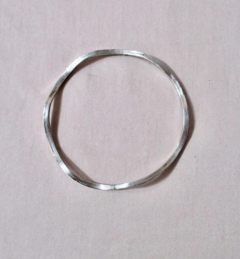 May include: A silver-colored, wavy bangle bracelet. The bracelet has a simple, minimalist design with a slightly irregular shape. The metal appears to be polished, reflecting light. The bracelet is a piece of jewelry.