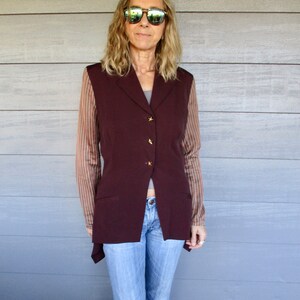 May include: A woman wearing a maroon waistcoat over a striped shirt, blue jeans, and sunglasses.