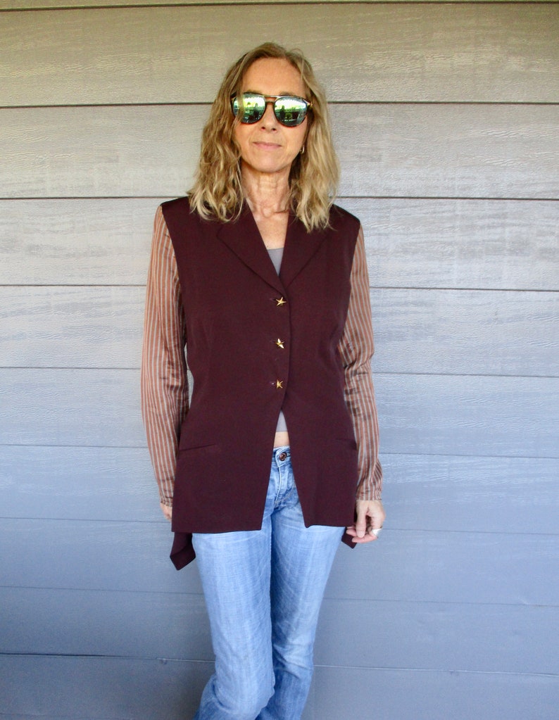May include: A woman wearing a maroon waistcoat over a striped shirt, blue jeans, and sunglasses.