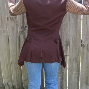 May include: A brown sleeveless waistcoat with a long, pointed hemline. The waistcoat has a collar and two pockets. The waistcoat is worn over a striped shirt and blue jeans.