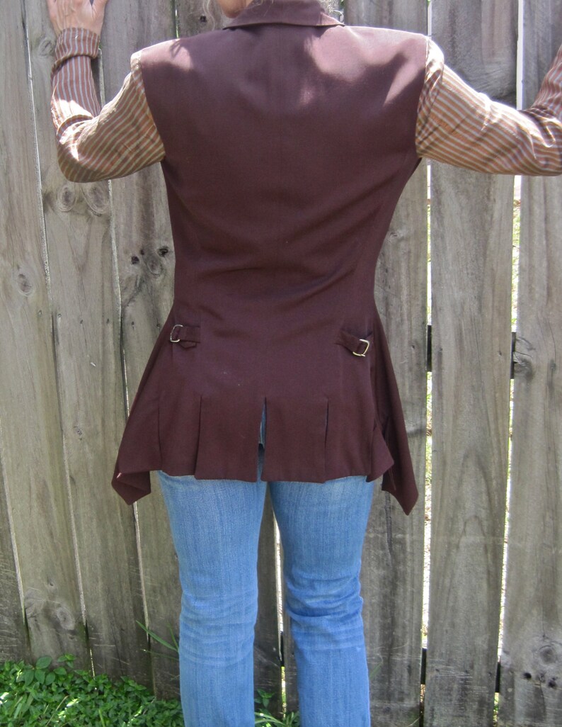 May include: A brown sleeveless waistcoat with a long, pointed hemline. The waistcoat has a collar and two pockets. The waistcoat is worn over a striped shirt and blue jeans.