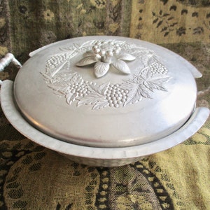May include: A silver aluminum serving dish with a lid. The lid has a floral design with grapes and leaves. The dish has two handles on the sides.