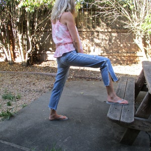 May include: A person wearing a pink and white tie-dye tank top and blue jeans with frayed hems. They are standing on a concrete surface with a wooden bench behind them.