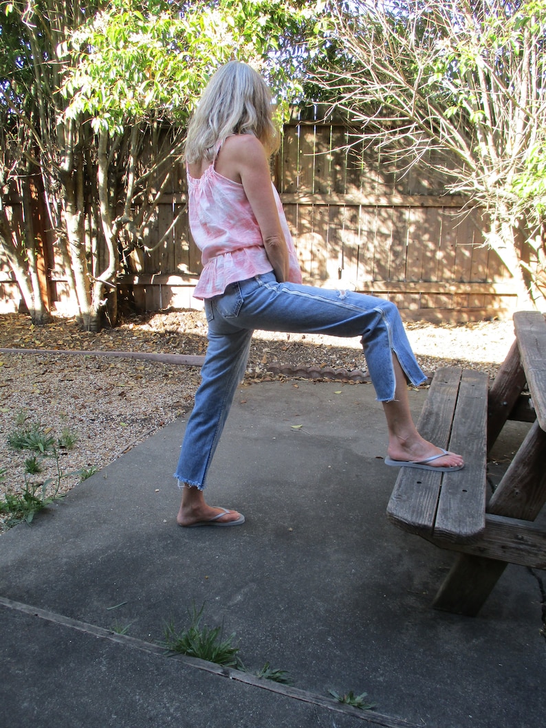 May include: A person wearing a pink and white tie-dye tank top and blue jeans with frayed hems. They are standing on a concrete surface with a wooden bench behind them.