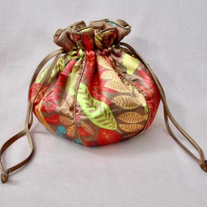 May include: A round drawstring pouch with a colorful leaf pattern in red, green, and brown. The pouch is made of a shiny fabric and has a brown drawstring closure. The overall design is a floral theme.