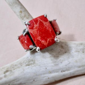 May include: A silver ring featuring three rectangular, vibrant red coral stones. The coral has a natural, textured pattern. The ring is set against a neutral background, highlighting the bold color and design of the jewelry.