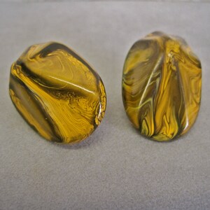 May include: Two oval-shaped earrings with a marbled design. The earrings feature a mix of yellow, brown, and olive green colors. The surface of the earrings is glossy and smooth, reflecting light.