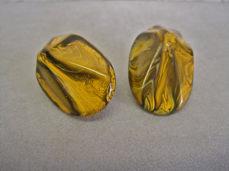 May include: Two oval-shaped earrings with a marbled design. The earrings feature a mix of yellow, brown, and olive green colors. The surface of the earrings is glossy and smooth, reflecting light.