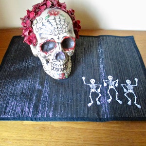 May include: A black and silver table runner with three white skeleton figures dancing on it. A white sugar skull with red flowers is on top of the runner.