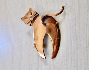 May include: A copper-colored cat brooch with a stylized design. The cat is facing left and has a long, thin tail.