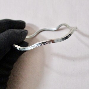 May include: A silver-colored, wavy bangle bracelet. The bracelet has a textured surface and a unique, organic shape. The design is simple yet elegant, suitable for everyday wear or special occasions. The bracelet is held by a gloved hand.