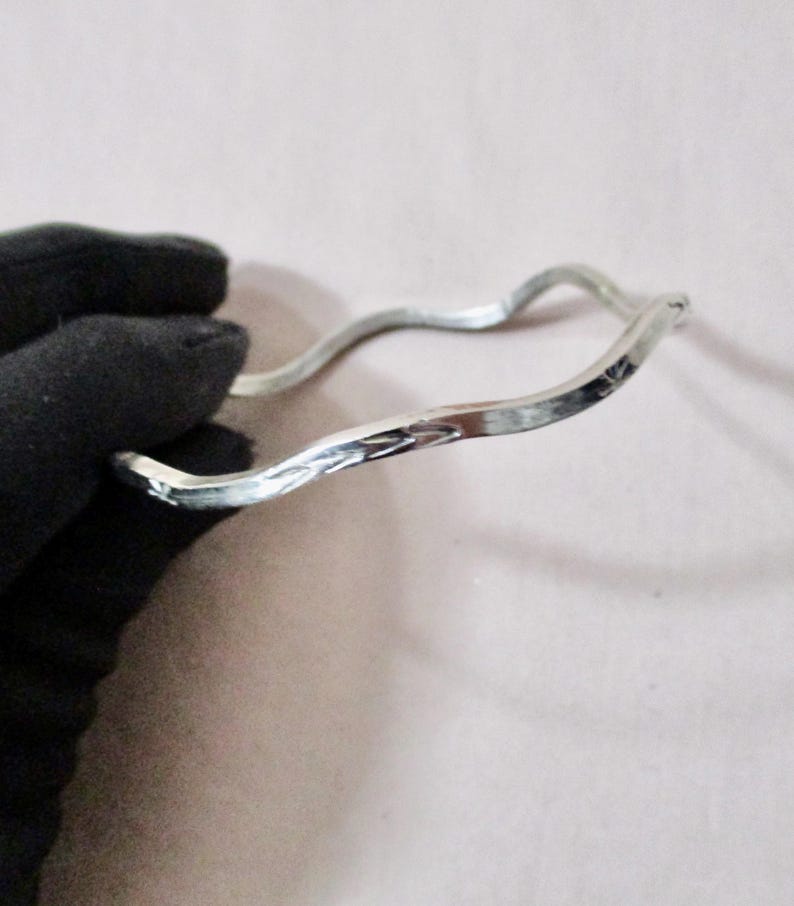 May include: A silver-colored, wavy bangle bracelet. The bracelet has a textured surface and a unique, organic shape. The design is simple yet elegant, suitable for everyday wear or special occasions. The bracelet is held by a gloved hand.