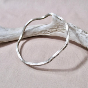 May include: A silver-colored, wavy bangle bracelet. The bracelet has a hammered texture and a unique, irregular shape. The bracelet is displayed on a light pink surface with a white antler in the background.