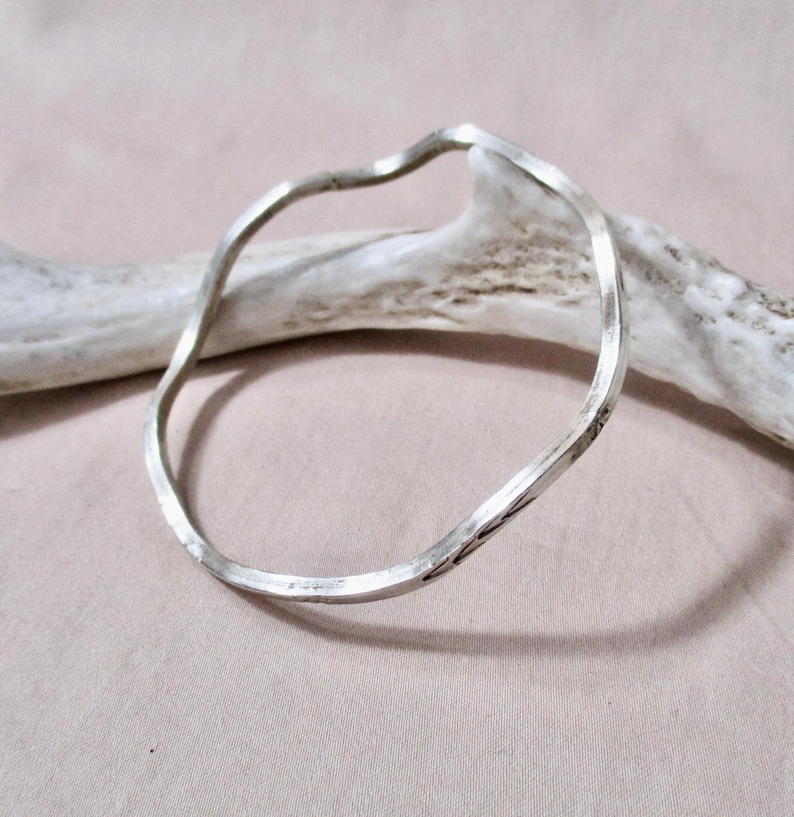 May include: A silver-colored, wavy bangle bracelet. The bracelet has a hammered texture and a unique, irregular shape. The bracelet is displayed on a light pink surface with a white antler in the background.