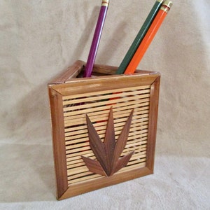 May include: A triangular bamboo pencil holder with a woven design and a leaf-shaped cutout. Three pencils are in the holder, one purple, one green, and one orange.