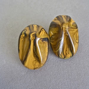 May include: Two oval-shaped earrings with a marbled pattern in shades of gold and brown. The earrings have a glossy finish and are set against a gray background. These earrings are a unique accessory.