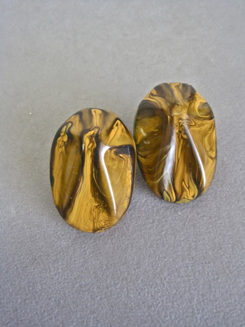 May include: Two oval-shaped earrings with a marbled pattern in shades of gold and brown. The earrings have a glossy finish and are set against a gray background. These earrings are a unique accessory.