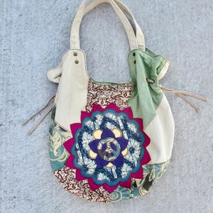May include: A hobo-style shoulder bag featuring a patchwork design of cream, sage green, and teal fabric panels. The center displays a large, multi-layered floral applique in magenta, blue, and purple, centered with a circular peace sign emblem.