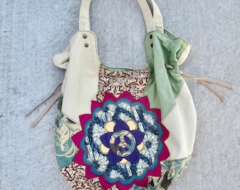 May include: A hobo-style shoulder bag featuring a patchwork design of cream, sage green, and patterned fabric panels. The center displays a large, scalloped magenta applique surrounding a circular blue and purple mandala with a peace sign emblem.