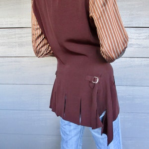 May include: A brown waistcoat with a long, pointed tail and a silver buckle. The waistcoat has a pleated design on the back and is worn over a striped shirt.