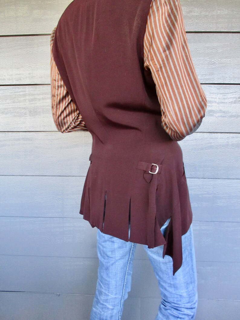 May include: A brown waistcoat with a long, pointed tail and a silver buckle. The waistcoat has a pleated design on the back and is worn over a striped shirt.