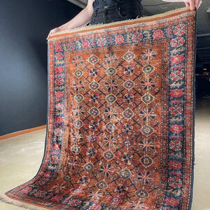 Könnte beinhalten: Ein rechteckiger, vintage Teppich mit einem komplizierten geometrischen Muster. Der Teppich hat ein warmes, rötlich-braunes Mittelfeld mit einem sich wiederholenden Design aus blauen und goldenen Formen. Der Rand ist dunkelblau mit floralen Motiven und der Teppich hat einen Fransenrand.