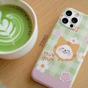 Pode incluir: Capa de telemóvel verde claro com um design de gato de desenho animado, flores brancas e o texto "MUV". A capa tem uma base rosa e um padrão xadrez verde. Uma chávena de matcha latte verde está no fundo.