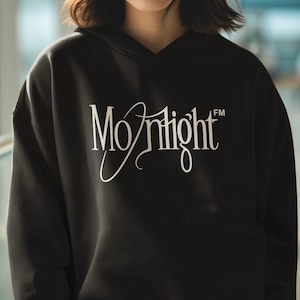 Op de afbeelding: Zwarte hoodie met het woord "Moonlight" in witte sierlijke letters op de borst. De sweatshirt heeft een capuchon met trekkoord en lange mouwen. De stijl is casual en comfortabel.