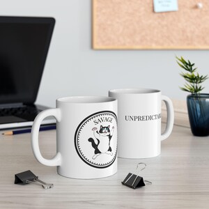 May include: Two white ceramic mugs. One mug features a black and white cat illustration with the word "SAVAGE" above it. The other mug has the word "UNPREDICTABLE" printed on it. Both mugs have a white handle.