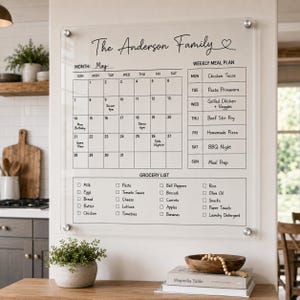 May include: A clear acrylic wall-mounted calendar board featuring the text The Anderson Family, a monthly calendar grid for May, a weekly meal plan section, and a grocery list with checkboxes. The board is secured with four silver metal standoffs.