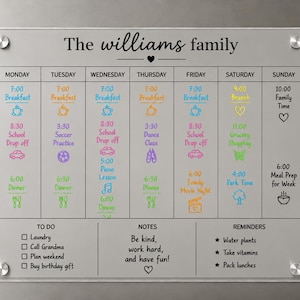 May include: A clear acrylic family calendar with the text "The williams family." The calendar is divided into days of the week with scheduled activities, reminders, and a to-do list. It is mounted on a metallic surface with silver screws.