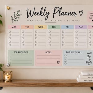 May include: A clear acrylic weekly planner board featuring black text that reads Weekly Planner, Plan, Focus, Achieve, Be Proud, Good Things Take Time, Top Priorities, Notes, This Week I Will, and Small Steps Big Results with pastel colored columns.