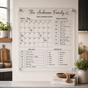 May include: A clear acrylic wall-mounted family command center board featuring a monthly calendar, weekly schedule, meal plan, and grocery list. The board is printed with black text including The Anderson Family, May, and various tasks.