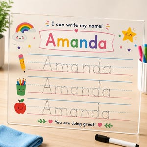 May include: A clear acrylic writing practice board featuring the text I can write my name! and the name Amanda in colorful letters. It includes three lines of dotted text for tracing, decorated with a rainbow, a star, a pencil, and an apple.