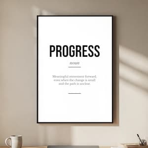 Puede incluir: Un cuadro negro con la palabra «PROGRESS» en negrita y su definición debajo. El cuadro está colgado sobre un escritorio de madera con un portátil, una taza y material de oficina.