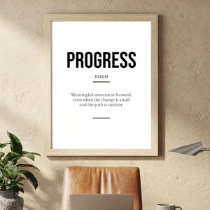 Può includere: Una stampa incorniciata con la parola "PROGRESS" in grassetto nero e la sua definizione sottostante. La stampa è sopra una scrivania con un laptop, una tazza e una pianta in vaso. È visibile anche una lampada da scrivania argentata.